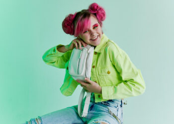 A person with vibrant pink hair styled in buns smiles while holding a small white bag. Their bright green jacket and ripped jeans evoke the playful spirit of 90s outfits. The background is a light, pastel green, enhancing their nostalgic vibe. | FintechZoom