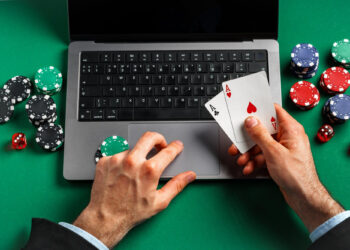 A person holding two aces, one of spades and one of hearts, in front of a laptop on a green table. Poker chips and dice are scattered around as the player uses their left hand on the laptop's trackpad, immersed in an online poker game. | FintechZoom