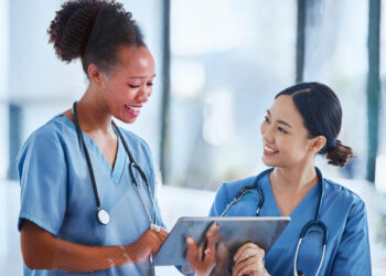 Two healthcare professionals are interacting. One, a woman with curly hair wearing blue scrubs and a stethoscope, is showing something on a tablet to her colleague, a woman with straight hair also in blue scrubs and a stethoscope. Both are smiling and engaged in discussing cost containment strategies. | FintechZoom