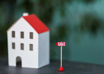 A small model of a white house with a red roof is placed on a dark surface, with a red sign that reads "SALE" positioned next to it. Promoting the idea to "Buy and Sell" while staying in your home, the background is blurred with green tones, hinting at tranquility and growth. | FintechZoom