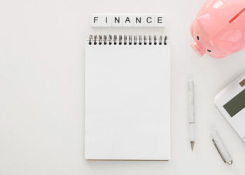 An open blank notebook is placed on a white surface. Above the notebook, the word "FINANCE" is spelled out with letter tiles. To the right, there is a pink piggy bank, a white calculator, a pen, and a USB flash drive—essential tools for managing paycheck to paycheck living. | FintechZoom