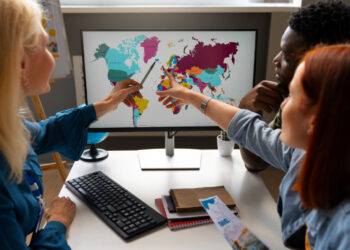 Three people sit around a desk, pointing at a world map displayed on a computer monitor. The desk is cluttered with a keyboard, notebooks, and travel brochures. It appears they are discussing travel plans or geography, possibly considering foreign exchange risk as they review the various countries in different colors. | FintechZoom