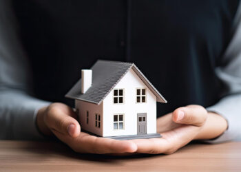 A person is holding a small model of a two-story house with both hands placed underneath it, representing a new mortgage. The house has a grey roof and white walls, and the person's torso is visible, dressed in a dark outfit with a light-colored shirt underneath. | FintechZoom