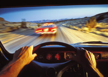 View from inside a speeding vehicle following a car with flashing lights on a desert road. The driver's hands grip the steering wheel, and the speedometer is illuminated. The surrounding landscape consists of desert terrain and distant mountains, setting the proximate cause for this high-speed pursuit. | FintechZoom