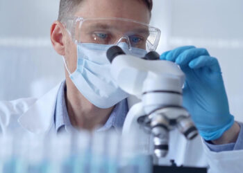 A scientist wearing safety goggles, a mask, and gloves is looking through a microscope in a laboratory. Various test tubes are visible in the foreground. The setting appears to be clean and sterile, indicating a controlled scientific environment studying antibiotic-resistant bacteria. | FintechZoom