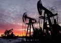 Silhouetted oil pump jacks operate against a dramatic sunrise or sunset sky with clouds. The scene captures several pump jacks in motion, indicating an active oil field where fluctuating oil prices drive continued activity. The ground is slightly snow-covered, suggesting a cold climate. | FintechZoom