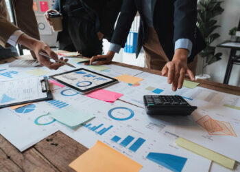 A group of people in business attire are gathered around a table covered with charts, graphs, and documents. One person is using a calculator, while another points at a tablet displaying a graph. Colored sticky notes adorn various documents as they engage in detailed financial management discussions. | FintechZoom