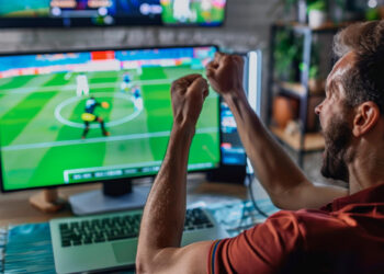 A man wearing a red shirt is sitting at a desk, enthusiastically cheering with raised fists while watching a soccer match on a computer monitor. The screen, tuned to a Sports Betting TV Channel, displays a dynamic in-game moment with players and a soccer field. | FintechZoom