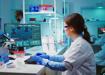 Scientists in lab coats and blue gloves work in a modern laboratory. A woman in the foreground analyzes data on a computer screen displaying cell images related to antibiotic-resistant infections. Test tubes, microscopes, and lab equipment are visible. Other scientists work diligently in the background. | FintechZoom