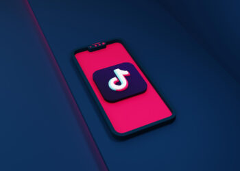 A smartphone with a pink screen displays the TikTok logo, a white musical note on a black background, in the center. The phone is set against a dark blue, geometric background with contrasting pink light reflections—perfect for attracting new TikTok followers. | FintechZoom
