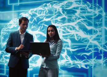 Two professionals, one man and one woman, collaborate while looking at a laptop and a tablet in front of a digital backdrop depicting a blue, illuminated brain with neural connections, symbolizing AI for Business and technology. | FintechZoom
