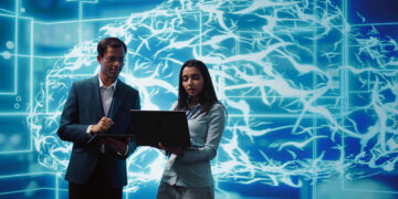 Two professionals, one man and one woman, collaborate while looking at a laptop and a tablet in front of a digital backdrop depicting a blue, illuminated brain with neural connections, symbolizing AI for Business and technology. | FintechZoom