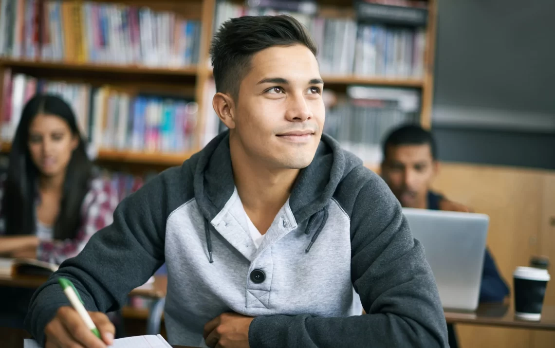 A young man in a gray hoodie sits at a desk in a library, holding a pen and writing on paper. He gazes off to the side with a thoughtful expression, perhaps contemplating his future. Bookshelves are visible in the background, and other students studying nearby might be considering their own Discover Student Loans options. | FintechZoom