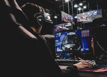 A person wearing headphones is seated in front of a gaming computer with a headset microphone. The computer screen displays a multiplayer video game. Overhead, illuminated gaming banners and screens are visible, suggesting a lively gaming event or tournament where the best VPN for gaming could be in use. | FintechZoom