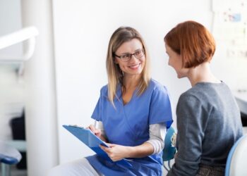 A healthcare professional in blue scrubs and glasses smiles while holding a clipboard and speaking with a woman who has short red hair and is wearing a grey top, possibly discussing options like a small business loan for her dentist practice. They are in a medical setting with bright lighting. | FintechZoom