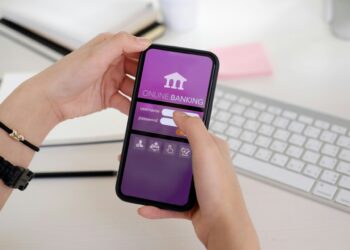 Two hands hold a smartphone displaying a fintech banking login screen with fields for username and password. A computer keyboard and notebook are visible in the background on a white desk. | FintechZoom