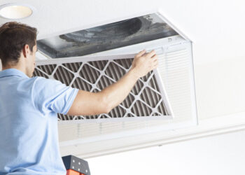 A person standing on a ladder and changing a 16x25x1 furnace air filter in a ceiling vent. They are wearing a light blue polo shirt and have dark hair. The ceiling vent is open, and the person is inserting the new filter. A recessed ceiling light is visible nearby. | FintechZoom
