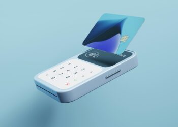 A sleek, modern fintech card reader with a small digital display and a numeric keypad is shown against a blue background. A blue credit card with a chip hovers above the reader, indicating contactless payment capability. | FintechZoom