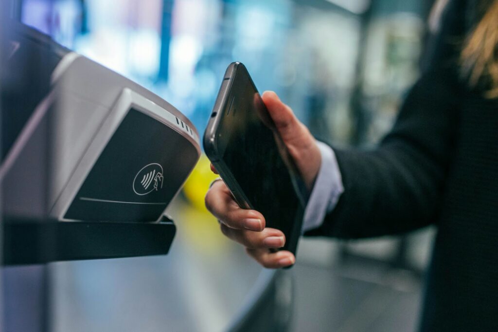 A person holding a smartphone near a contactless payment terminal, indicating a contactless transaction. The individual is wearing a dark sleeve, and the background appears to be an indoor setting with blurred elements. | FintechZoom