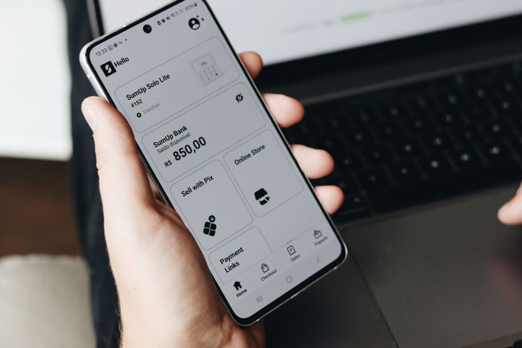 A person holds a smartphone displaying a banking app interface. The screen shows account details, including a balance of R$ 850.00, and options like "Sell with Pix," "Online Store," and "Payment Links." A laptop keyboard is visible in the background. | FintechZoom