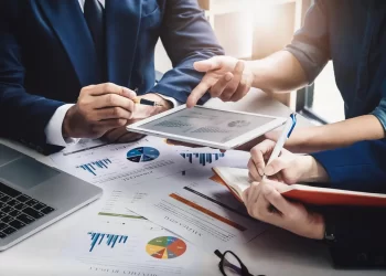 Three individuals in business attire are engaged in a discussion around a table filled with financial charts and graphs. One person, representing FintechZoom.com Business, is holding a tablet displaying more data, while another takes notes in a notebook. A laptop is also visible on the table. | FintechZoom