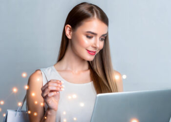 A woman with long hair is holding a shopping bag and using a laptop. She is smiling and wearing a sleeveless top. Bright spots of light surround her, creating a dynamic and modern atmosphere. | FintechZoom