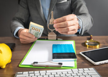 A person in a suit holds house-shaped keychains over a desk with a notepad, calculator, magnifying glass, smartphone, and a stack of cash. A yellow piggy bank is also visible. The scene suggests a real estate or financial setting. | FintechZoom