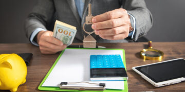 A person in a suit holds house-shaped keychains over a desk with a notepad, calculator, magnifying glass, smartphone, and a stack of cash. A yellow piggy bank is also visible. The scene suggests a real estate or financial setting. | FintechZoom