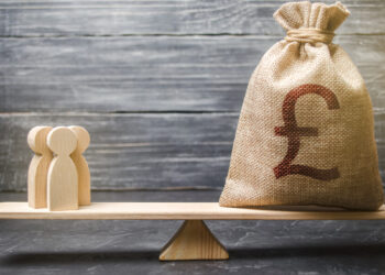 A burlap sack with a large British pound symbol is balanced on a wooden seesaw opposite small wooden figurines, suggesting a comparison between people and money. The background is a weathered, gray wooden surface. | FintechZoom
