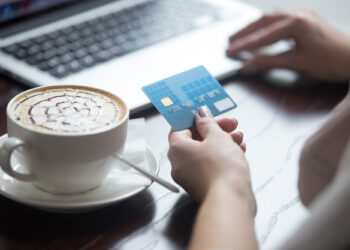 A person holds a blue Visa card, simplifying online transactions while using a laptop. A cappuccino with latte art sits on the table beside them. | FintechZoom