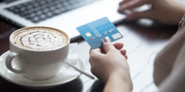 A person holds a blue Visa card, simplifying online transactions while using a laptop. A cappuccino with latte art sits on the table beside them. | FintechZoom