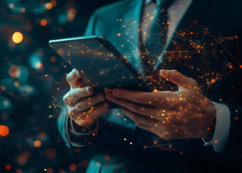 A person in a suit holds a tablet, surrounded by digital, glowing network connections. The background is dark, highlighting the vibrant, orange light trails that illustrate the power of modern technology and connectivity in the realm of digital business. | FintechZoom