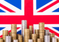 Stacks of various coins, symbolizing growth amid UK inflation, are arranged in front of a large Union Jack flag. The coins are grouped in different heights, suggesting wealth, with the flag serving as a patriotic backdrop. | FintechZoom
