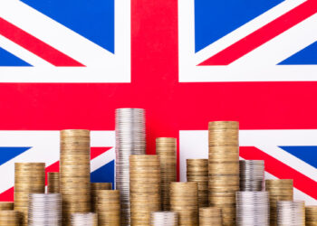 Stacks of various coins, symbolizing growth amid UK inflation, are arranged in front of a large Union Jack flag. The coins are grouped in different heights, suggesting wealth, with the flag serving as a patriotic backdrop. | FintechZoom