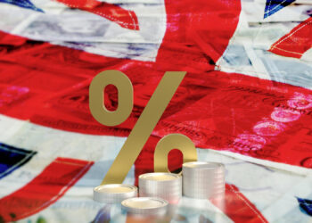 A large golden percentage symbol stands in front of stacks of coins, overlaying a Union Jack flag and various British banknotes. The composition suggests themes of finance and economic interest rates in the UK context. | FintechZoom