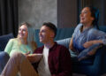 Three friends are sitting on a couch, smiling and watching Netflix. One person holds a bowl of popcorn, adding to the cozy living room setting as they appear relaxed and entertained. | FintechZoom