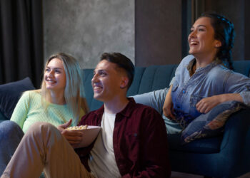 Three friends are sitting on a couch, smiling and watching Netflix. One person holds a bowl of popcorn, adding to the cozy living room setting as they appear relaxed and entertained. | FintechZoom