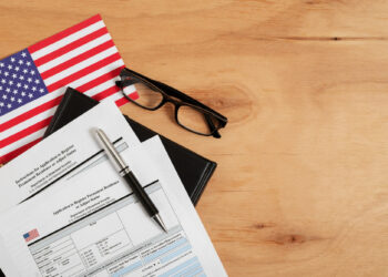 A wooden surface with printed tax forms, a black pen, reading glasses, and a small American flag. The items are arranged neatly, suggesting a setting for work or study related to finance or taxation. | FintechZoom