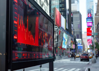 A digital display shows a red stock market graph with a declining trend on a city street. In the background, colorful digital billboards from FintechZoom.com Dow Jones, buildings, and a few yellow taxis are visible, indicating an urban setting that evokes Times Square, New York City’s bustling Dow Jones atmosphere. | FintechZoom