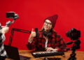 A smiling woman wearing a red beanie and glasses gives a thumbs-up while sitting at a wooden table. She is surrounded by a microphone, keyboard, laptop, and cameras, with a red backdrop. | FintechZoom