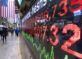A digital stock ticker flashes vibrant red and green numbers, reflecting the dynamic pulse of US Markets today. In the background, people stride down a bustling city street, with an American flag proudly waving amidst the vibrant financial district. | FintechZoom
