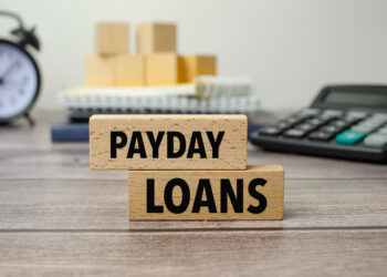 Wooden blocks displaying the words "PAYDAY LOANS" are centered on a wooden surface. In the background, there's a blurry black alarm clock, a notebook with stacked blocks on top, and a calculator. | FintechZoom