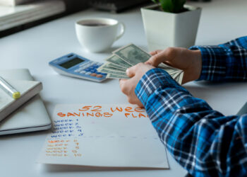 A person in a blue plaid shirt counts dollar bills at a desk, seemingly browsing FintechZoom.com Money for money-saving tips. Next to them is a calculator, a cup of coffee, a pen, a notebook, and a sheet of paper labeled "52 Week Savings Plan," with weeks 1 to 10 listed but empty. | FintechZoom