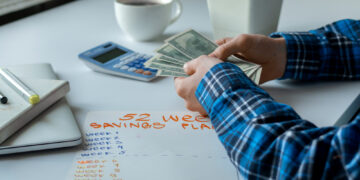 A person in a blue plaid shirt counts dollar bills at a desk, seemingly browsing FintechZoom.com Money for money-saving tips. Next to them is a calculator, a cup of coffee, a pen, a notebook, and a sheet of paper labeled "52 Week Savings Plan," with weeks 1 to 10 listed but empty. | FintechZoom