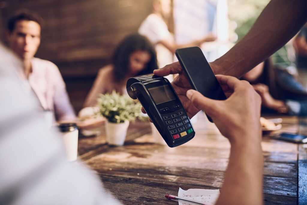 A person uses a smartphone for mobile payment through a card reader at a wooden table, with a group of people sitting in the background and a potted plant nearby. | FintechZoom