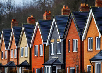 A row of modern, colorful two-story houses with gabled roofs and brick exteriors. Each house features white-framed windows and neatly arranged chimneys. Trees without leaves can be seen in the background under a cloudy sky. | FintechZoom