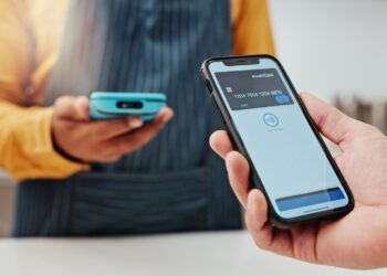 A person in a yellow shirt holds a blue card reader, while another uses a smartphone to make a contactless payment, showcasing how tech is changing payments and personal finance. The payment app is visible on the phone's screen, illustrating seamless and modern transactions. | FintechZoom