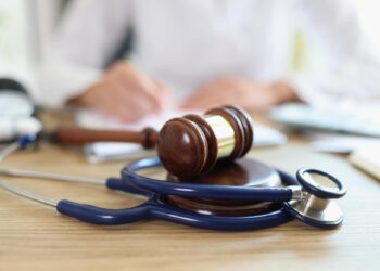A gavel and a stethoscope rest on a wooden desk, symbolizing the intersection of law and medicine. In the background, slightly out of focus, a person in a white coat writes—a scene reflecting the crucial role of personal injury lawyers in navigating these fields. | FintechZoom