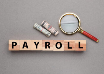 Wooden blocks spelling "PAYROLL" are arranged in a row. Beside them, a rolled-up stack of dollar bills and a magnifying glass are placed on a gray surface, suggesting financial scrutiny or analysis. | FintechZoom