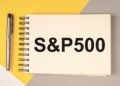 A notebook with "S&P500" written on it is placed on a yellow and gray background. A silver pen is positioned on the left side of the notebook. | FintechZoom
